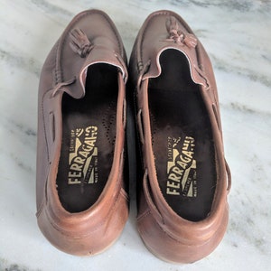 May include: A pair of brown leather loafers with tassels. The loafers have a gold label that reads "FERRAGAMO" and "FIRENZE" with the text "MADE IN ITALY" underneath.