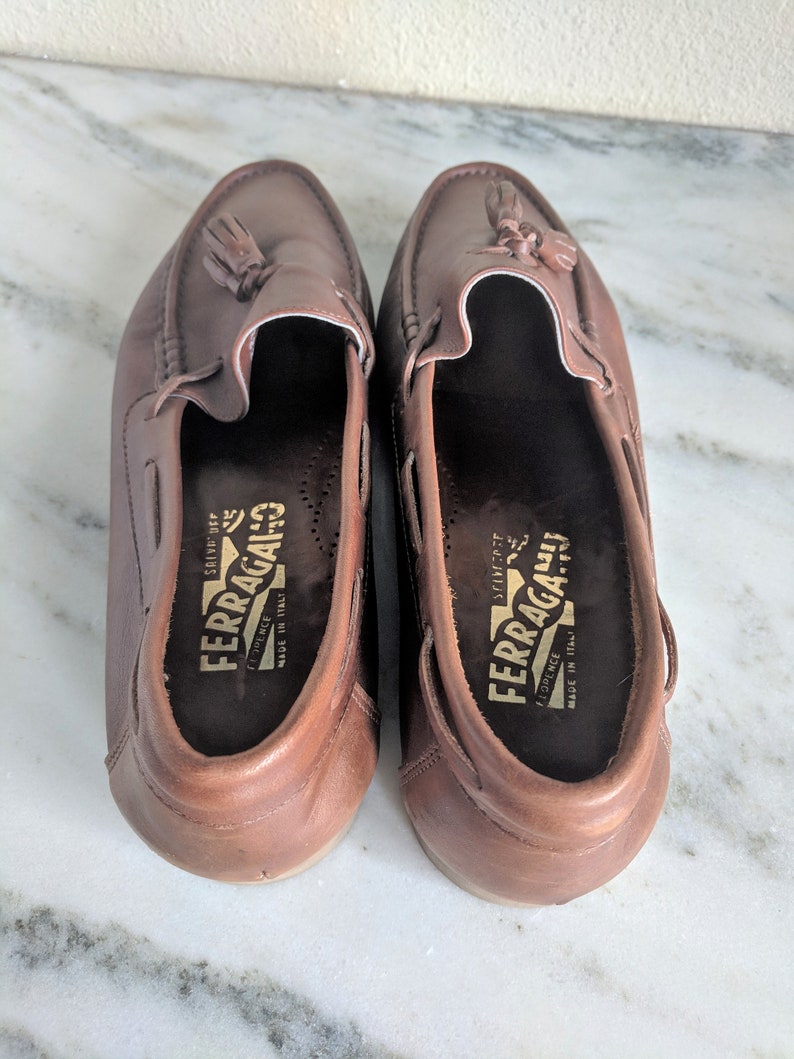 May include: A pair of brown leather loafers with tassels. The loafers have a gold label that reads "FERRAGAMO" and "FIRENZE" with the text "MADE IN ITALY" underneath.