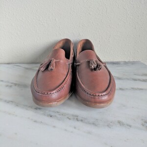 May include: A pair of brown leather tassel loafers with a stitched edge. The loafers are on a white marble surface.
