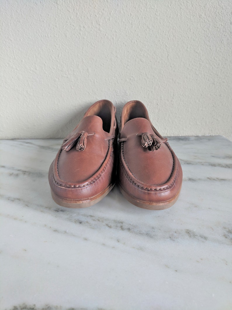 May include: A pair of brown leather tassel loafers with a stitched edge. The loafers are on a white marble surface.