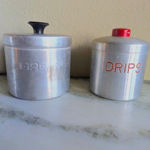 以下が含まれることがあります： 2つのヴィンテージアルミニウム製キッチンキャニスター。1つのキャニスターは黒いハンドルと前面にエンボス加工された「GREASE」の文字があります。もう1つのキャニスターは赤い蓋と赤い文字で「DRIPS」の文字があります。