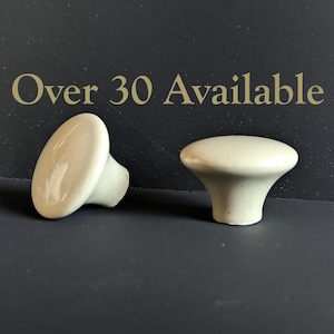 May include: Two off-white ceramic cabinet knobs with a rounded top and a flared base. The knobs are displayed against a dark gray background, with the text "Over 30 Available" in gold lettering.