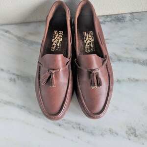 May include: A pair of brown leather loafers with tassels. The loafers have the brand name "Ferragamo" printed on the inside.
