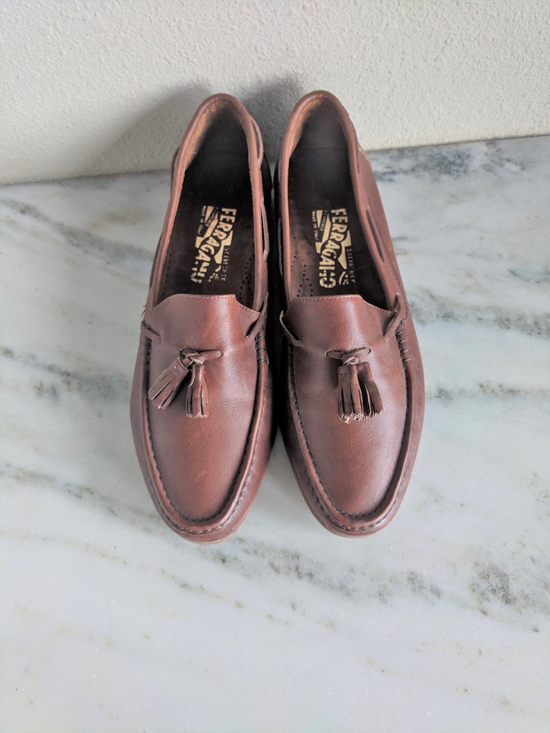 May include: A pair of brown leather loafers with tassels. The loafers have the brand name "Ferragamo" printed on the inside.
