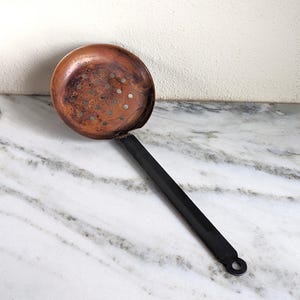 May include: A copper slotted spoon with a black handle. The spoon has a round bowl with holes in it. The handle is long and thin.