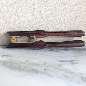 May include: A pair of wooden cheese knives with a dark reddish-brown finish. The knives have a unique design with a gold-colored accent in the center. The handles are rounded, and the blades are angled. The knives are displayed on a marble surface.