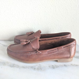 May include: Brown leather tassel loafers with a boat shoe style. The loafers have a brown leather sole and a tassel detail on the front.