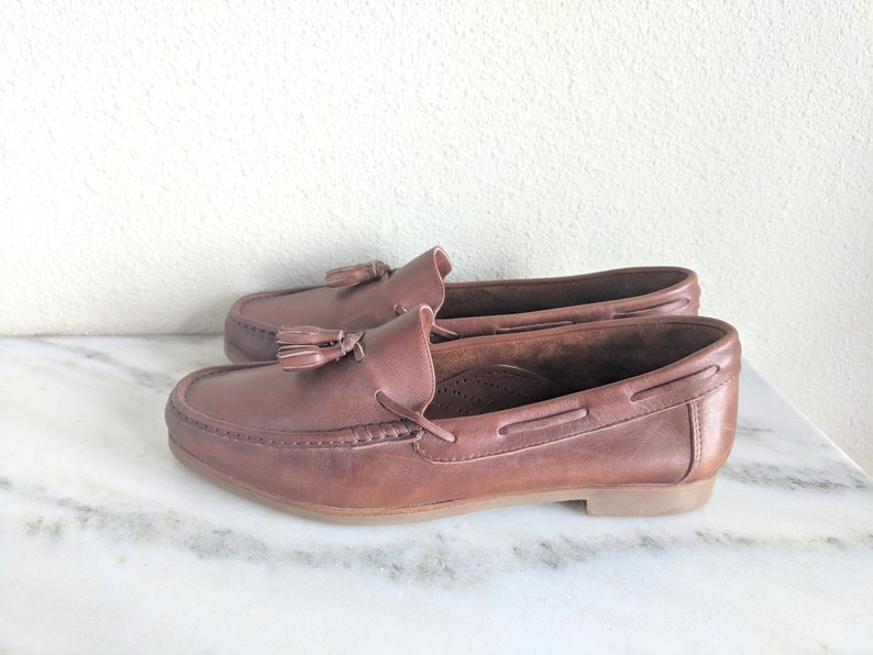May include: Brown leather tassel loafers with a boat shoe style. The loafers have a brown leather sole and a tassel detail on the front.