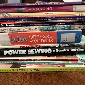 May include: A stack of sewing and quilting books, including titles like "Creative Serging," "Power Sewing," and "One-Yard Wonders." The books are in various colors and sizes, with visible text on the spines. The stack is on a brown surface.