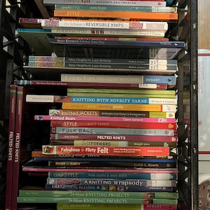 May include: A black plastic crate filled with a large collection of stacked knitting and craft books. The spines display various titles including Socks Socks Socks, Reversible Knits, Knitted Bears, Felted Knits, and 24-Hour Knitting Projects.
