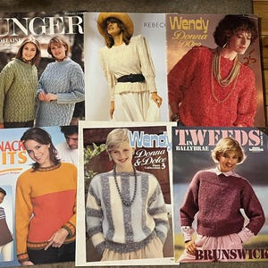 May include: A collection of vintage knitting pattern magazines featuring women's sweaters. The magazines showcase various sweater designs in different colors and styles, including cable knit and striped patterns. The covers display models wearing the knitted garments.