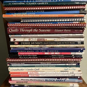 May include: A stack of quilting books with titles like "Quilts Through the Seasons" and "501 Quilt Blocks." The books are in various colors, including red, blue, and white, and are stacked on top of each other. Some books have spiral bindings.