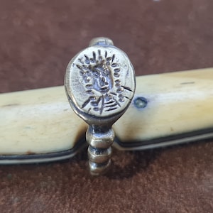 May include: A silver ring with an oval-shaped top featuring a detailed engraved design. The ring has a beaded band.