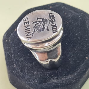 K&ouml;nnte beinhalten: Ein silberner Ring mit einer ovalen Oberseite, die einen stilisierten L&ouml;wen und den Text "GEMINA LEG-XIII" zeigt.