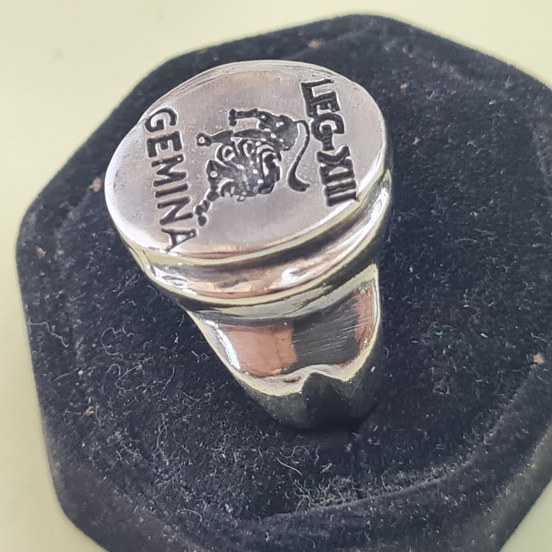 K&ouml;nnte beinhalten: Ein silberner Ring mit einer ovalen Oberseite, die einen stilisierten L&ouml;wen und den Text "GEMINA LEG-XIII" zeigt.
