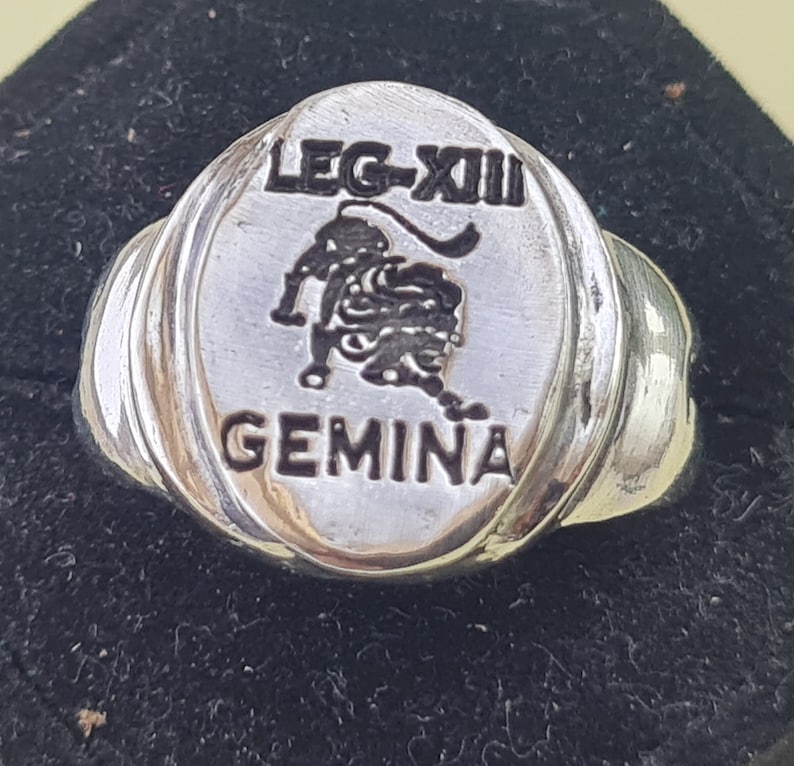 K&ouml;nnte beinhalten: Ein silberner Ring mit einer ovalen Oberseite, die ein Schwarzwei&szlig;bild des Sternzeichen Zwillinge, den Text "LEG-XII" und das Wort "GEMINA" zeigt.