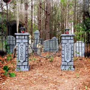 May include: A spooky Halloween cemetery scene. Two stone pillars with "HAUNTED" and "CEMETERY" text flank a wrought iron gate. The scene includes tombstones, a chain, and a red lantern. The ground is covered in brown leaves, with a forest backdrop.
