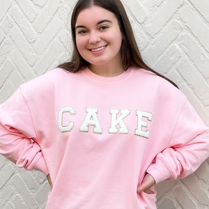 Könnte beinhalten: Ein rosa Sweatshirt mit dem Wort "CAKE" in weißen Blockbuchstaben auf der Vorderseite. Das Sweatshirt wird von einer Person getragen, die vor einer weißen Backsteinwand steht.