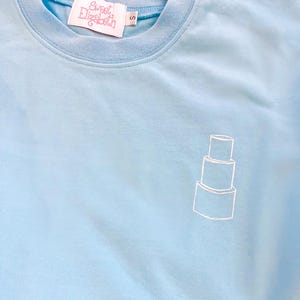 Könnte beinhalten: Ein hellblaues T-Shirt mit einem weißen Umriss eines dreistöckigen Kuchens auf der Vorderseite. Das Shirt hat ein Etikett mit der Aufschrift "Sweet Elizabeth" und "S" für die Größe.
