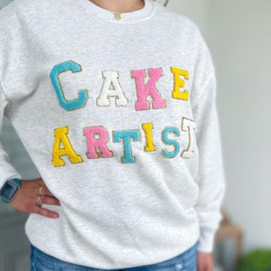 Könnte beinhalten: Hellgraues Sweatshirt mit dem Schriftzug "CAKE ARTIST" in bunten, gestickten Buchstaben. Die Buchstaben sind goldfarben umrandet, mit einem blauen "C", einem rosa "K", einem gelben "E", einem rosa "A", einem gelben "R", einem blauen "T", einem gelben "I" und einem blauen "S".