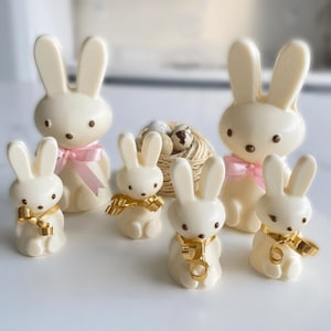 May include: Five white chocolate bunny figurines with pink and gold ribbon accents. The bunnies are arranged in a group on a white surface.
