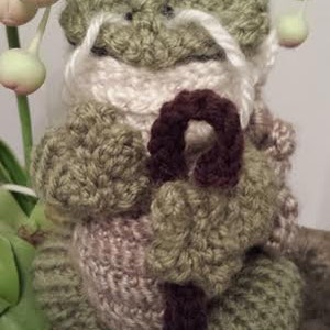 May include: A crocheted frog wearing a white scarf and holding a brown stick. The frog is green and has white whiskers.