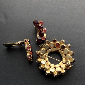 May include: Three vintage brooches featuring red and pink rhinestones. One brooch is a circle with a pin back, another is a cluster of stones with a clip back, and the third is a single row of stones with a clip back.
