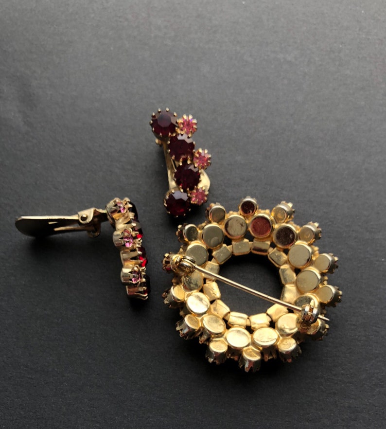 May include: Three vintage brooches featuring red and pink rhinestones. One brooch is a circle with a pin back, another is a cluster of stones with a clip back, and the third is a single row of stones with a clip back.
