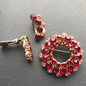 May include: A set of vintage jewellery featuring a large red and pink rhinestone brooch in the shape of a circle and two matching earrings. The brooch is made of gold-toned metal and has a pin back.