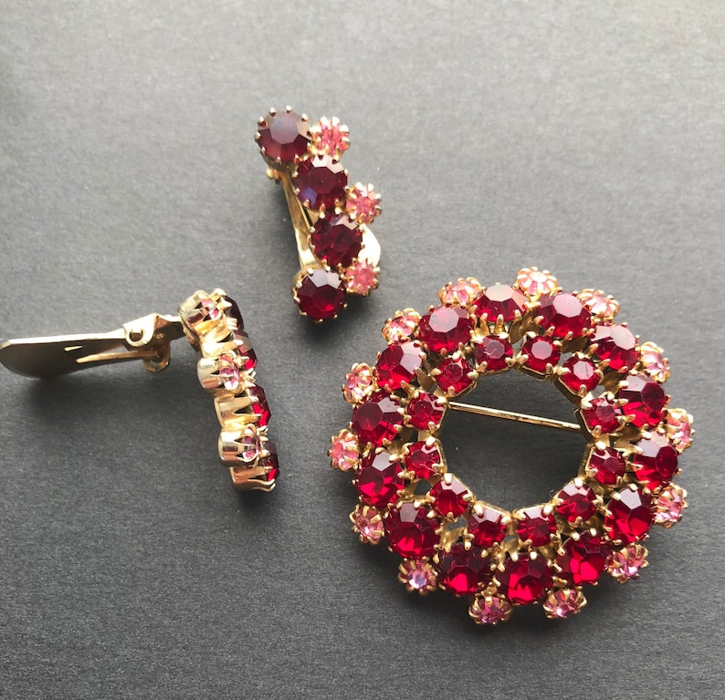 May include: A set of vintage jewellery featuring a large red and pink rhinestone brooch in the shape of a circle and two matching earrings. The brooch is made of gold-toned metal and has a pin back.