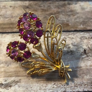 Könnte beinhalten: Eine goldfarbene Brosche mit zwei violetten Blütenbüscheln und einem dekorativen goldenen Blattdesign.