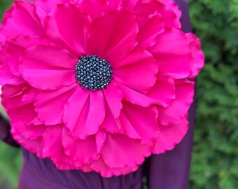 May include: A large, bright pink fabric flower with a dark blue jeweled center. The flower is worn as a brooch on a purple dress.