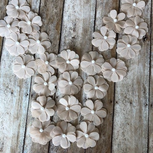 May include: A collection of beige fabric flowers with white pearl centers. The flowers are arranged in a V-shape on a rustic wooden background.