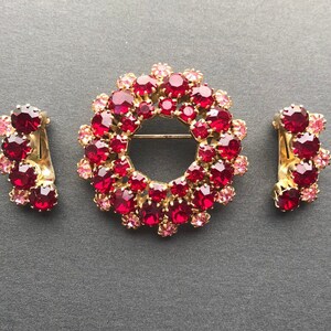 May include: A set of three gold-tone jewellery pieces with red and pink rhinestones. The set includes a brooch in the shape of a circle, and two earrings.