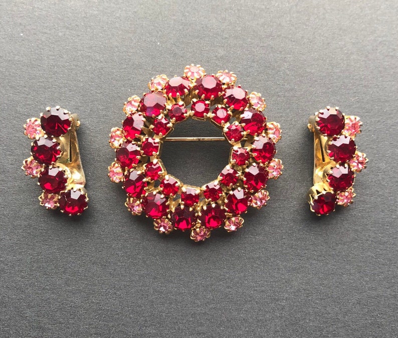 May include: A set of three gold-tone jewellery pieces with red and pink rhinestones. The set includes a brooch in the shape of a circle, and two earrings.