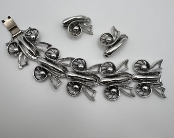 Op de afbeelding: Een zilverkleurige armband met een sluiting en een bijpassend paar oorbellen. De armband is gemaakt van een reeks gekoppelde, sierlijke, zilverkleurige strikken met een kleine, ronde, zilverkleurige kraal in het midden van elke strik.