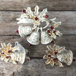 Könnte beinhalten: Ein Set vintage Weihnachts-Schmuck mit einer Brosche und Ohrringen. Die Brosche ist aus goldfarbenem Metall mit weißer Emaille-Oberfläche und zeigt eine Gruppe von Glocken mit Stechpalmenblättern und roten Strasssteinen. Die Ohrringe sind ein passendes Paar mit Glocken- und Stechpalmenmotiven.