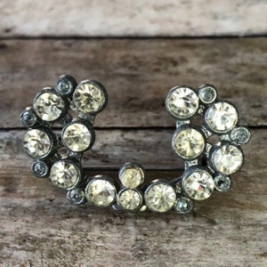 May include: A silver-toned brooch with a horseshoe shape, adorned with clear rhinestones.