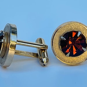 May include: A pair of gold cufflinks with a round, faceted, dark red gemstone in each. The cufflinks have a textured gold finish.