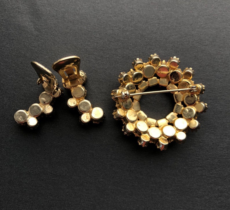 May include: A gold-tone brooch and a pair of earrings with a circular design of small, round, clear rhinestones.