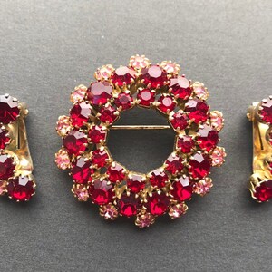 May include: A set of vintage jewellery featuring a large red and pink rhinestone brooch and a pair of matching earrings. The brooch is shaped like a circle and the earrings are shaped like a crescent moon.