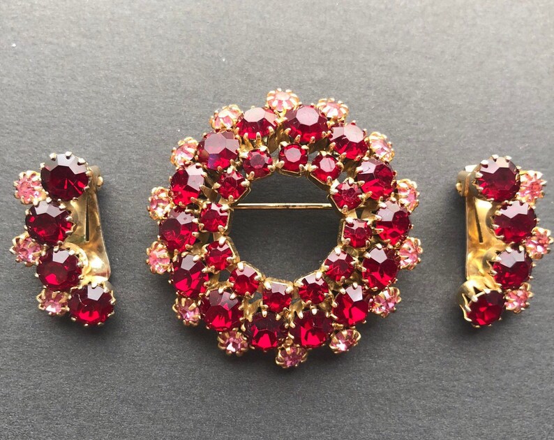 May include: A set of vintage jewellery featuring a large red and pink rhinestone brooch and a pair of matching earrings. The brooch is shaped like a circle and the earrings are shaped like a crescent moon.