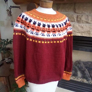 May include: A long-sleeved knit sweater in a deep maroon color featuring a round neckline. The yoke displays a fair isle pattern with alternating black cats and orange pumpkins, accented by white, yellow, and orange bands at the cuffs and collar.