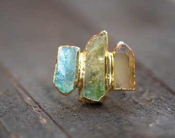 Könnte beinhalten: Ein goldener Ring mit drei verschiedenen Edelsteinen. Die Edelsteine sind hellblau, grün und hellbraun.