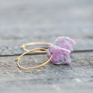 Puede incluir: Un par de pendientes de aro dorados con detalles de piedras preciosas rosas y blancas. Los pendientes están sobre una superficie de madera.