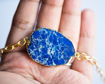 Puede incluir: Un brazalete de cadena dorada con un gran colgante de piedra de forma irregular, azul y blanco.