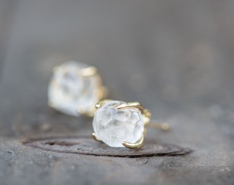 May include: Close-up of two clear, faceted crystal stud earrings with gold-colored prongs. The earrings are set against a blurred, neutral background, highlighting the sparkling crystals. The jewelry is designed to be worn as an accessory.