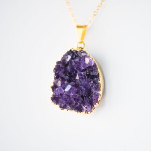 May include: A gold chain necklace with a pendant featuring a large amethyst crystal cluster. The crystal is a deep purple color and has a natural, rough texture.
