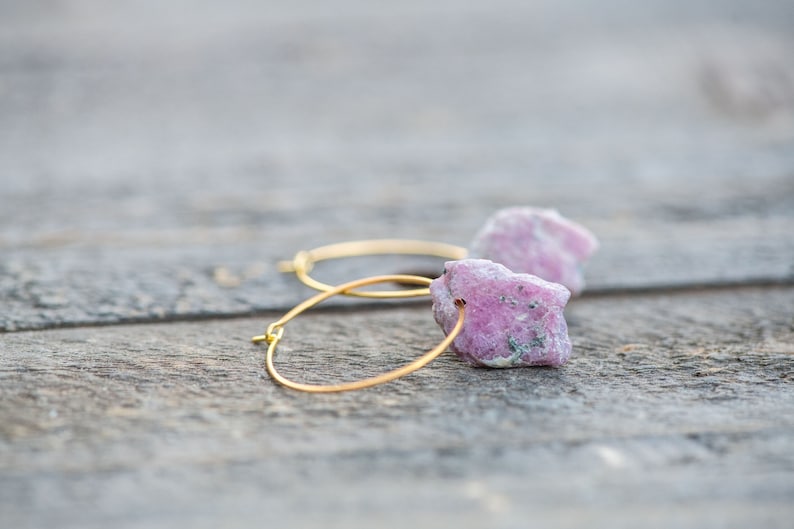 Peut inclure: Une paire de boucles d'oreilles cr&eacute;oles dor&eacute;es avec des accents de pierres pr&eacute;cieuses roses et blanches. Les boucles d'oreilles sont sur une surface en bois.