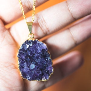 May include: A gold chain necklace with a large amethyst crystal pendant. The crystal is a cluster of purple crystals with a gold rim.
